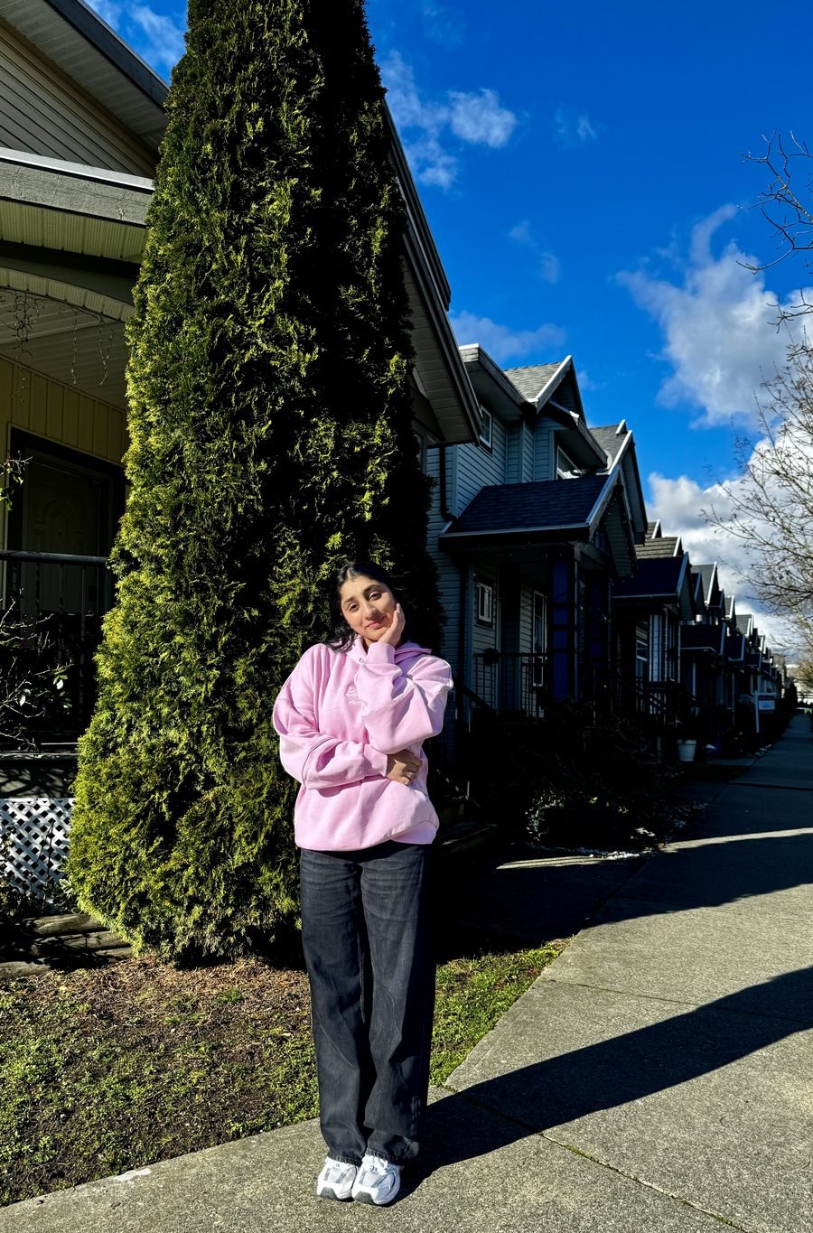 Harpreet smiling in a pink hoodie on a sunny street lined with trees